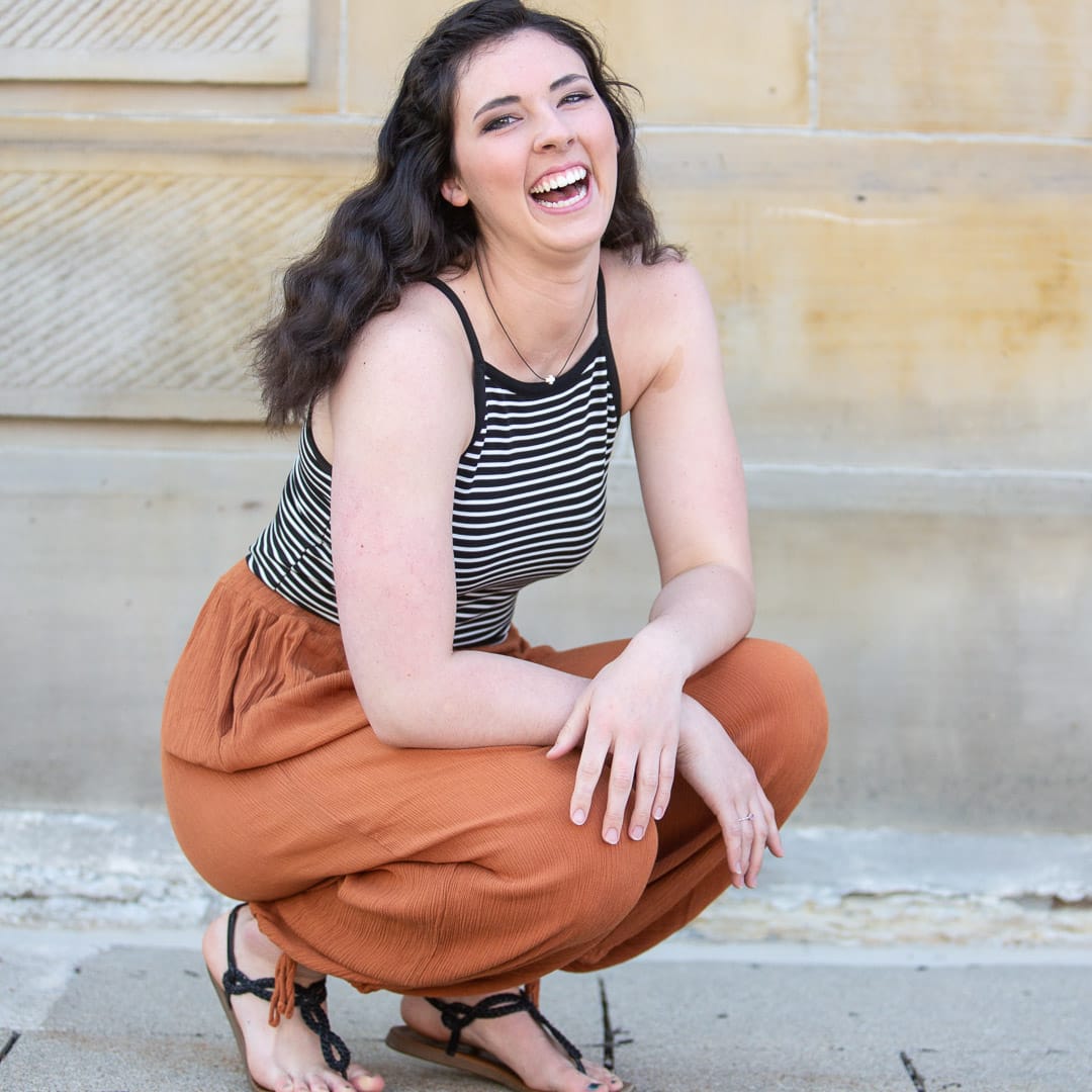 Noblesville Senior High School Student with long dark wavy hair wearing burnt orange flowing pants kneeling in front of the Hamilton County courthouse with a big laughing smile on her face. Credit: Studio Kate Portrait Design - Noblesville Senior Pictures