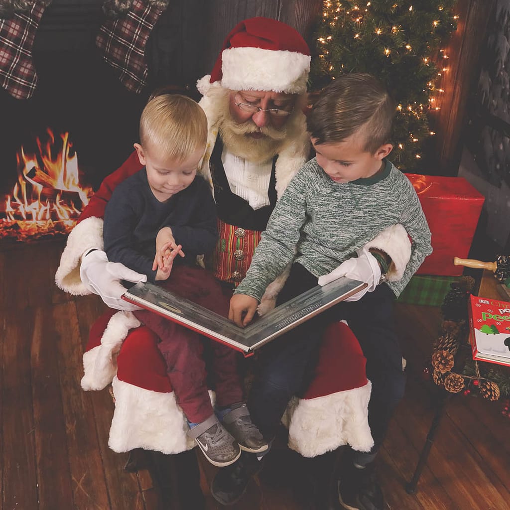 Two young boys sit on Santa's lap in front of a Christmas tree and fireplace. Santa is holding the Night Before Christmas book, while the older boy points to one of the pictures. Photographer Credit: Kate Plummer - Studio Kate Portrait Design - Noblesville Santa Experience