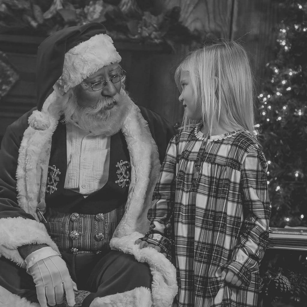 This black and white image features a young girl in her plaid pajamas standing next to Santa as she answer the traditional question of has she been a good girl this year. Photographer Credit: Kate Plummer - Studio Kate Portrait Design - Noblesville Santa Experience