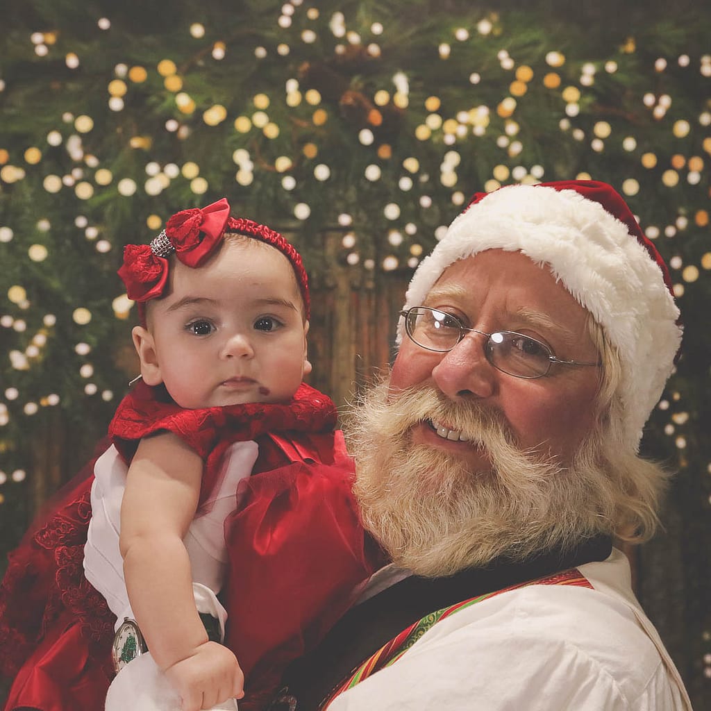 Santa holds a baby in a red satin dress with a matching hair bow. Photographer Credit: Kate Plummer - Studio Kate Portrait Design - Noblesville Santa Experience