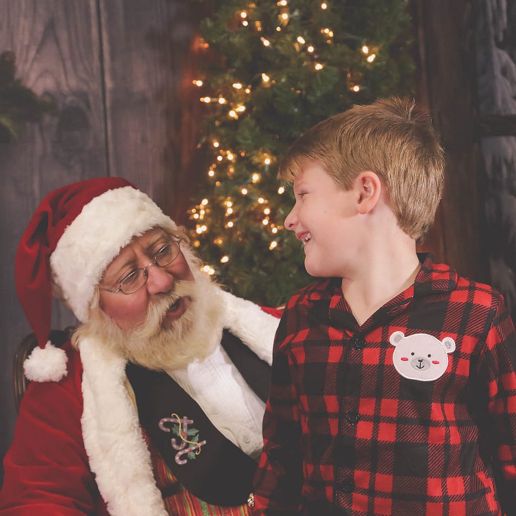 A young boy in red plaid pajamas looks back toward Santa Claus with a big smile. Photographer Credit: Kate Plummer - Studio Kate Portrait Design - Noblesville Santa Experience