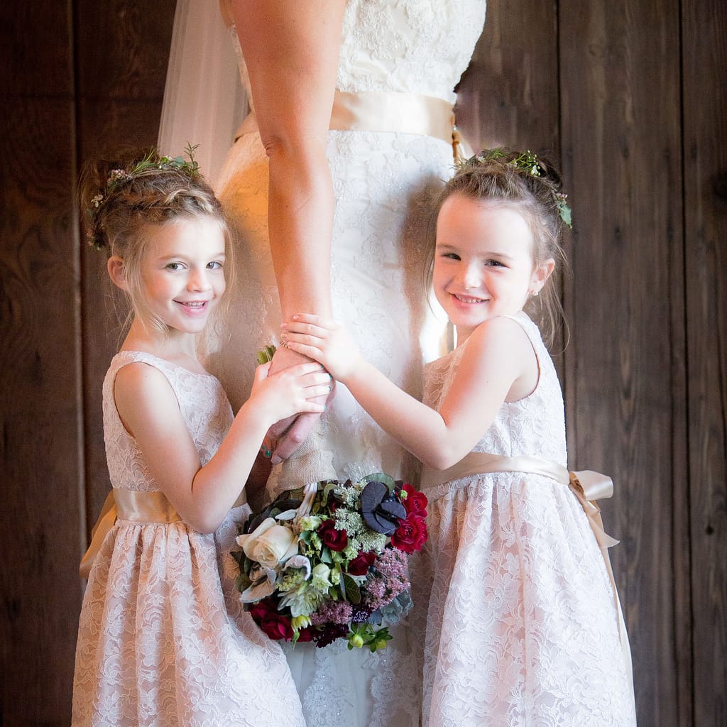 2 young girls in their junior bridesmaid dresses, holding the arm of the bride. This image is cropped so the bride is only visible from the waist down, while the young girls hold her closely as the bouquet falls between them Photo by Kate Plummer of Studio Kate Portrait Design.