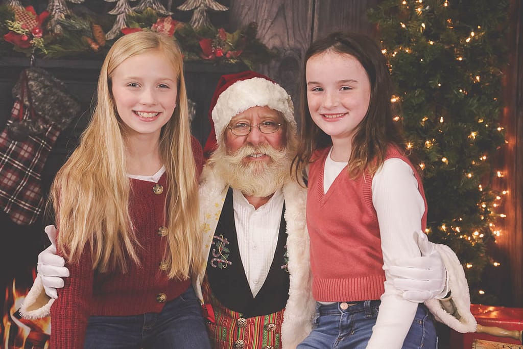 Two pre-teen girls sit on Santa's lap posing with smiles. Photographer Credit: Kate Plummer - Studio Kate Portrait Design - Noblesville Santa Experience