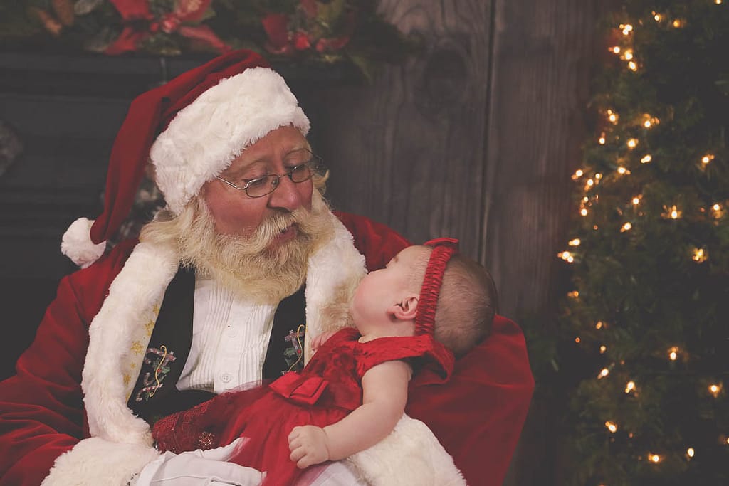 Santa holds a baby in a red satin dress with a matching hair bow. She's looking up at him with a smile. Photographer Credit: Kate Plummer - Studio Kate Portrait Design - Noblesville Santa Experience