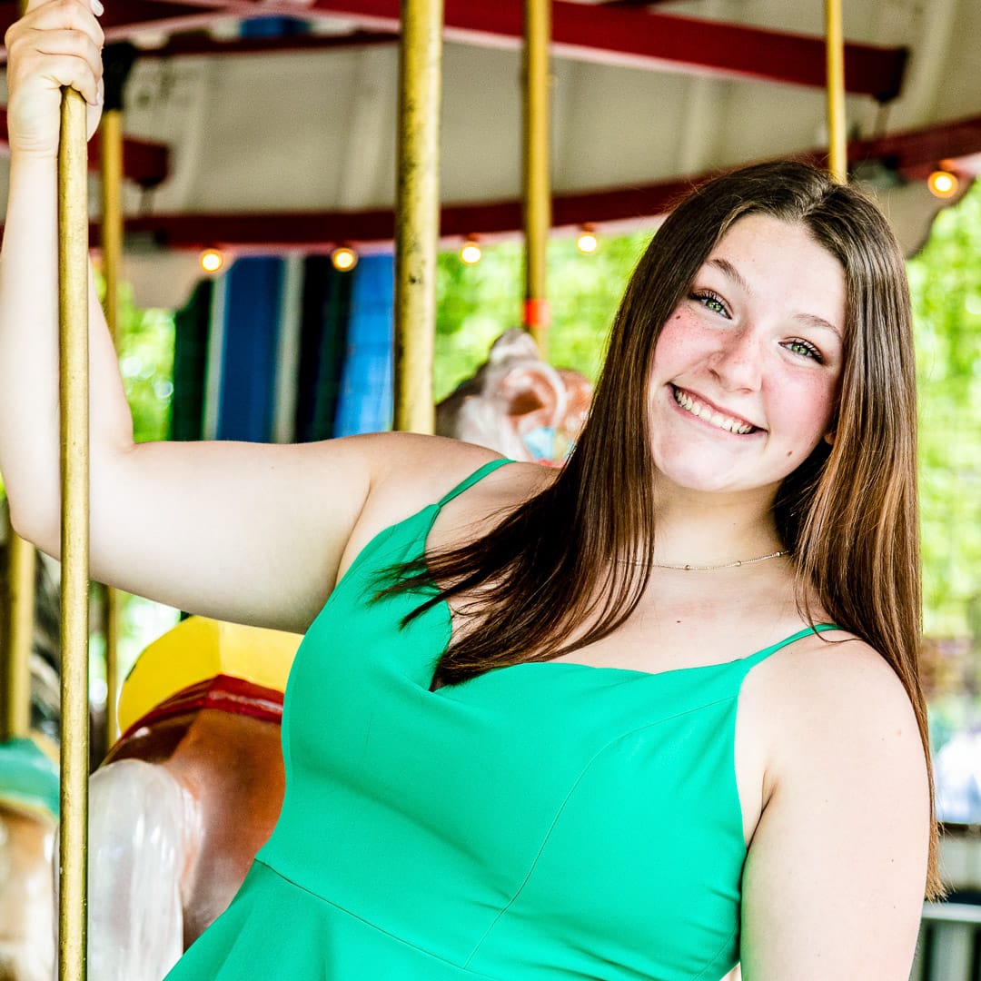 Noblesville Senior High School student is wearing a green summer dress while she stands on the edge of the carousel floor leaning out holding one of the classic polished brass bars.  You can see the carousel horses, classic roof lights, and the Forest Park trees softly focused in the background.