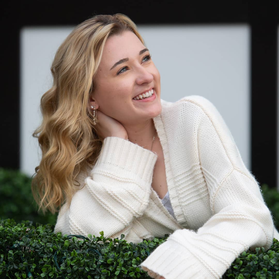 Noblesville Senior High School Student wearing a white sweater looking up and away with her hand on the back of her neck just behind her wavy blonde hair.  She’s leaning on some greenery in front of a black framed store window at Hamilton Town Center. Credit: Studio Kate Portrait Design - Noblesville Senior Pictures