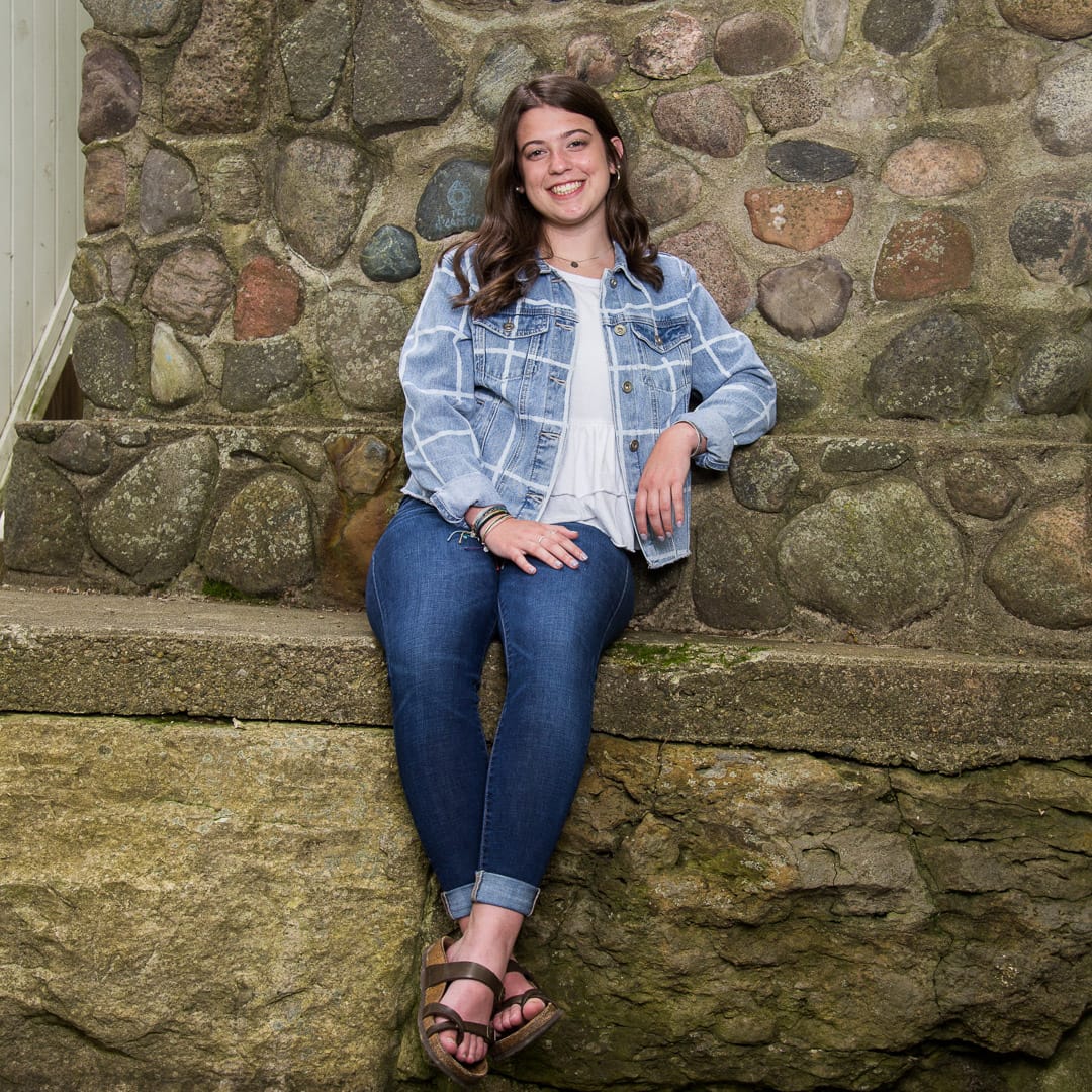 Noblesville Senior High School Student wearing a jean jacket sitting on the ledge of a rock wall built on one of the supports that holds up Potter’s Bridge. Credit: Studio Kate Portrait Design - Noblesville Senior Pictures