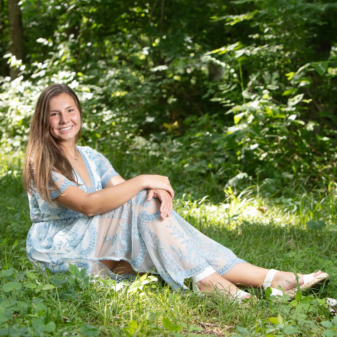 Noblesville Senior High School Student wearing a long blue dress sitting in the grass with spots of sunlight sneaking in along the White River Greenway. Credit: Studio Kate Portrait Design - Noblesville Senior Pictures