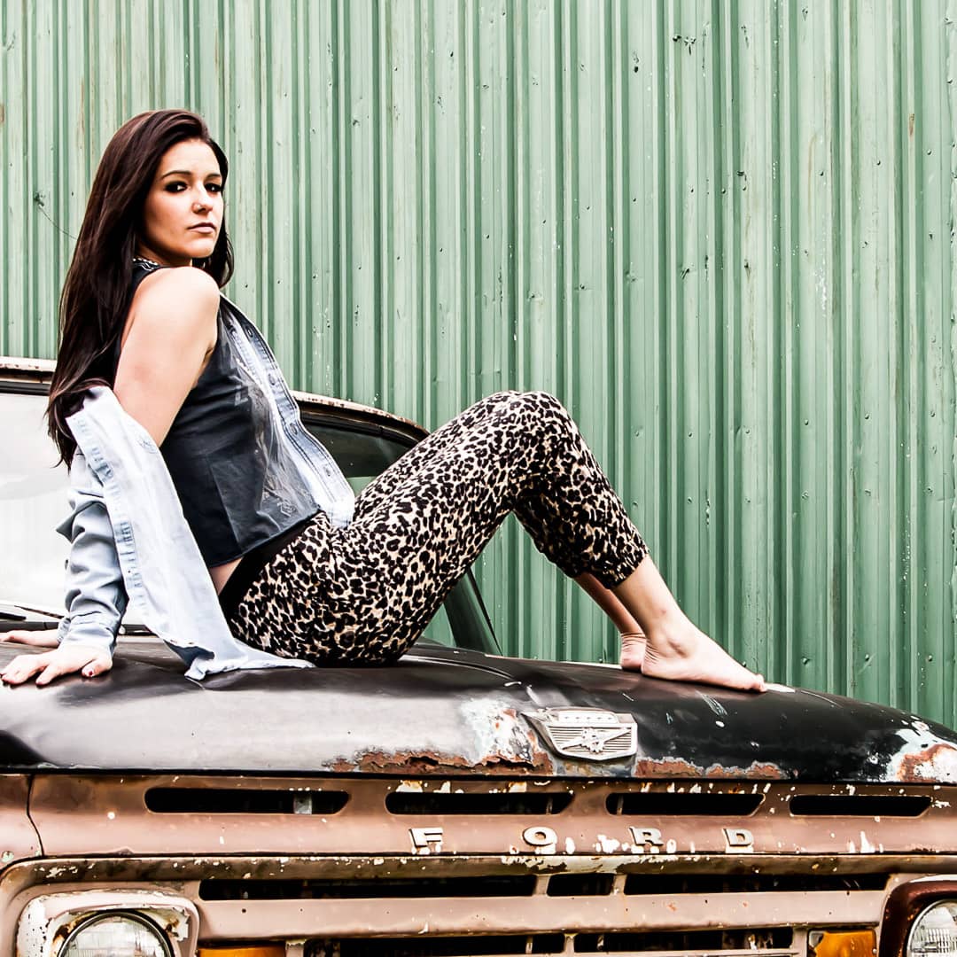 Noblesville Senior High School Student wearing favorite sleeveless concert t-shirt. She’s sitting on the hood of an rusty Ford truck, with her jacket hanging off her shoulder, in her bare feet. Credit: Studio Kate Portrait Design - Noblesville Senior Pictures