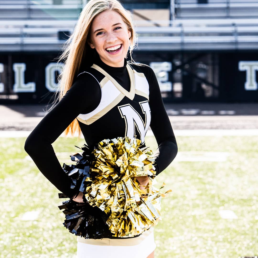 This Noblesville Senior High School cheerleader is standing on the NHS football field with her black and gold metallic pom poms at her hips.  The school’s “N” logo covers the torso  of her cheer uniform, and she has a big laughing smile on her face. 