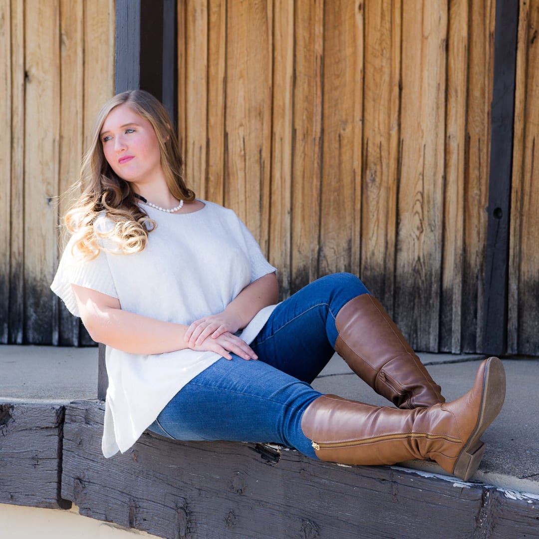 Noblesville Senior High School Student wearing jeans tucked into her knee high brown boots and a white flowing top.  She’s leaning against a post at Arbuckle’s Railroad Place. Credit: Studio Kate Portrait Design - Noblesville Senior Pictures