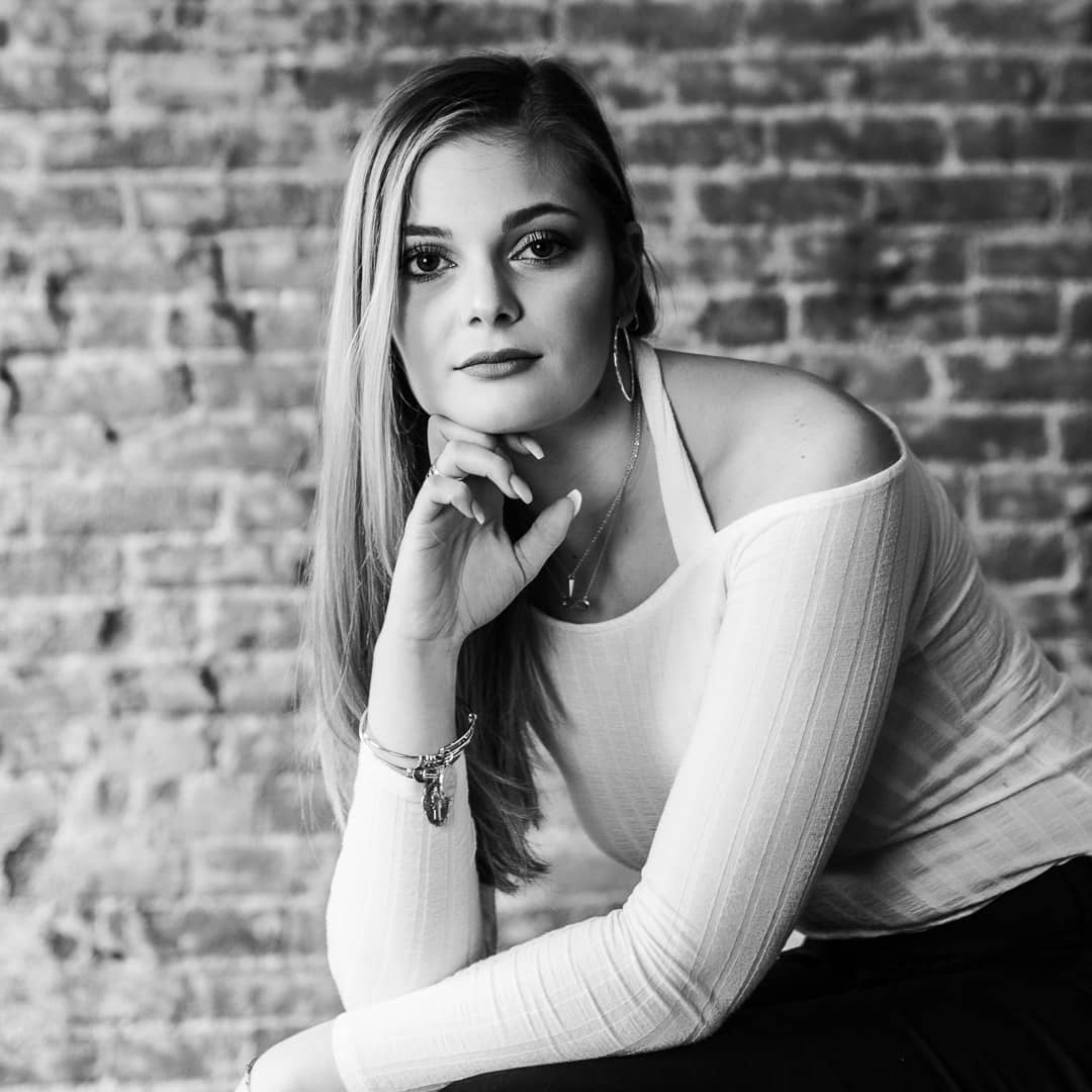 Noblesville Senior High School student wearing a while off the shoulder sweater and black pants.  She’s posed against the exposed brick studio wall in almost a “Thinker” statue position but looking directly at the camera with only the slightest hint of a smile.  Her shoulder length straight blonde hair is all pulled to one side and the dramatic light provides some darkened shadows on the right-side edges of her face.  Credit: Studio Kate Portrait Design - Noblesville Senior Pictures
