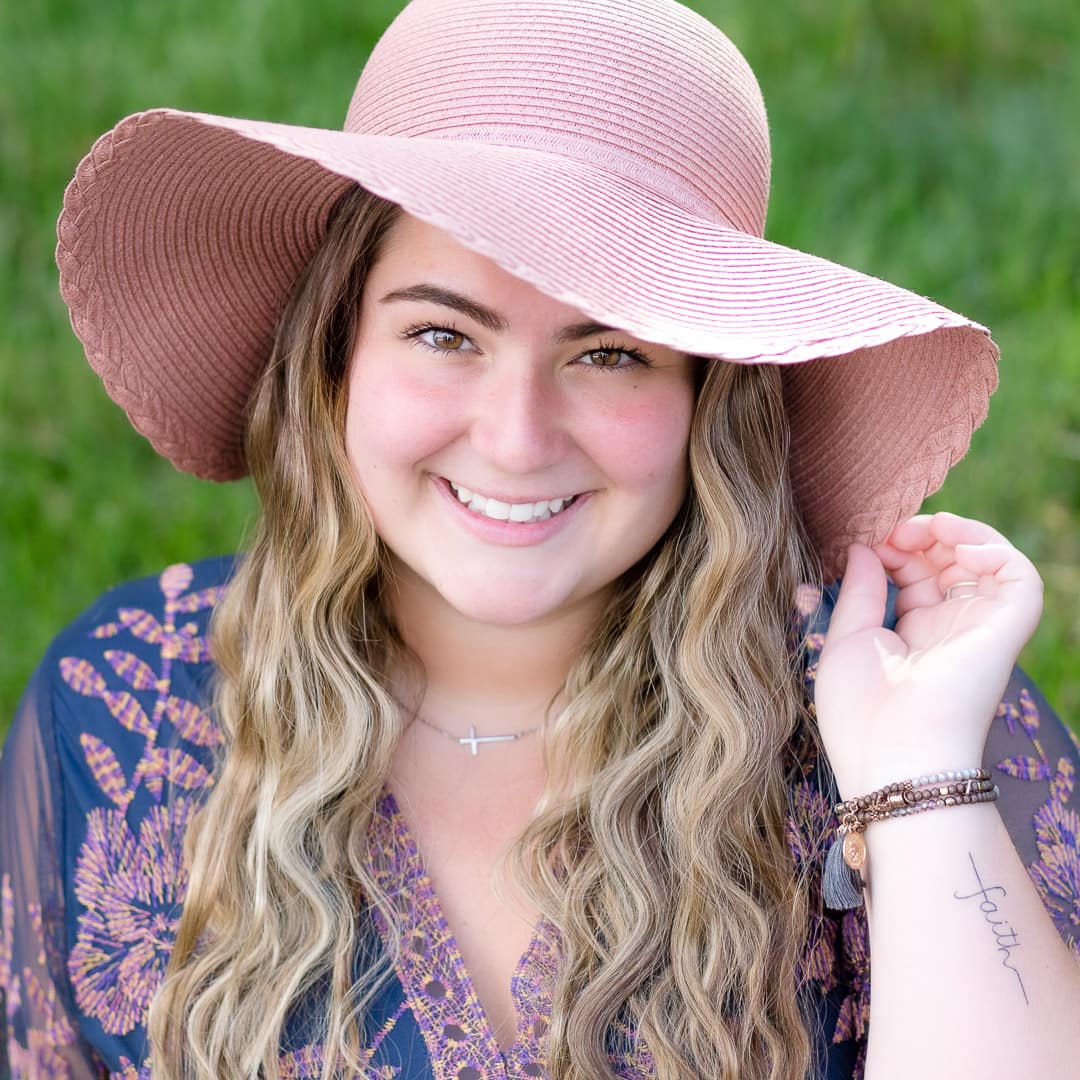 Noblesville Senior High School student wearing a big floppy sun hat.  Her long wavy hair is falling in front of her shoulders and her hand is gently pulling one side of the pink hat down. Her smile reaches from her mouth, up her cheeks, and all the way into her eyes.  Credit: Studio Kate Portrait Design - Noblesville Senior Pictures