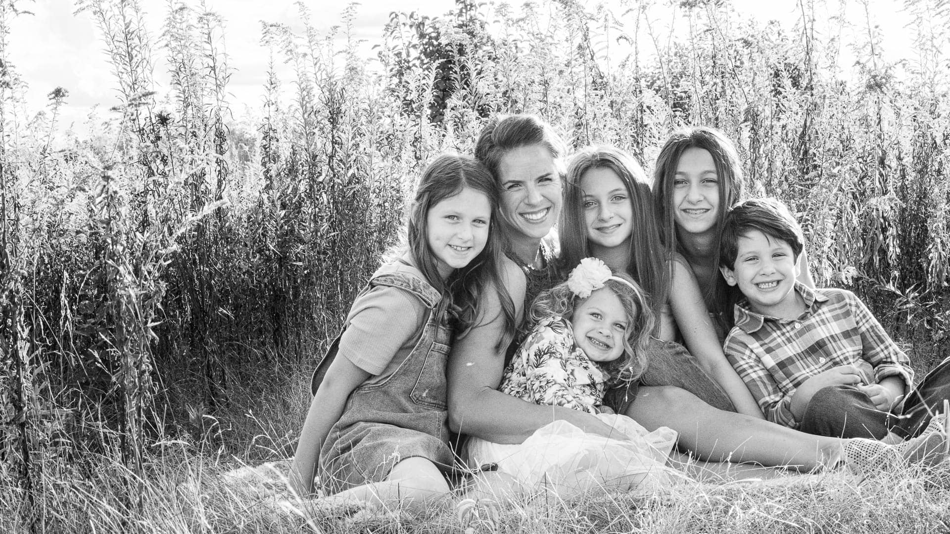 Beautiful Noblesville Family Portrait posed sitting together in a tall grassy field. This black and white image includes Mom, 4 daughters, and a son. There are smiles all around as the youngest two giggle. Photo Credit: Kate Plummer - Studio Kate Portrait Design - Noblesville Family Photographer