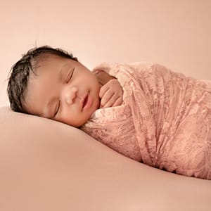 An infant with dark hair lies swaddled in a warm pink blanket with one tiny hand reaching out from the top. The child's head is resting on the soft continuous dune-like curve of the cashmere colored surface and matching background.