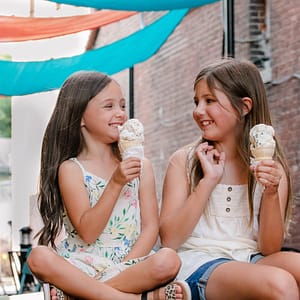 Two pre-teen sisters are sitting on a table in the alley North of Logan Street in the historic downtown square of Noblesville. They are looking at each other, giggling, with the teal and orange sun shades flying overhead. Photo Credit: Kate Plummer - Studio Kate Portrait Design - Noblesville Family Photographer