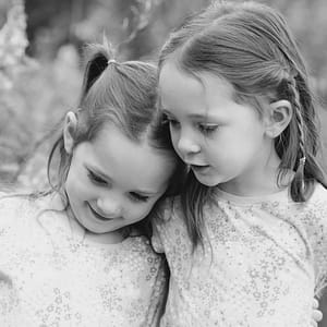 2 elementary aged sisters are posed together both looking down and sharing a moment. In the background you can see the soft focused trees that surround Potter's Bridge in Noblesville. Photo Credit: Kate Plummer - Studio Kate Portrait Design - Noblesville Family Portrait Photography