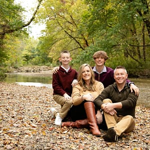 A young Noblesville family including Mom, Dad, and 2 teenage sons are posed on the rocky beach edge of the White River. It's a beautiful early fall setting with a few yellow and orange leaves among the rocks while the trees are still filled with green. Photo Credit: Kate Plummer - Studio Kate Portrait Design - Noblesville Family Pictures