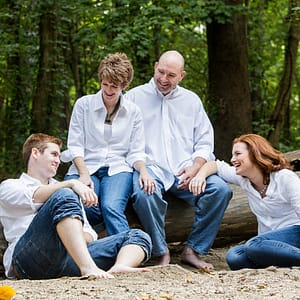 A family of 4 gathers on the beach of a local creek. Mom and Dad are sitting together on a large fallen tree, while the two college aged red headed siblings sit on the ground on each side of them. They are dressed in matching blue jeans and white button down shirts. Photo Credit: Kate Plummer - Studio Kate Portrait Design - Noblesville Family Photographer