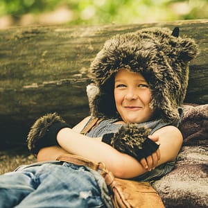 An elementary aged boy is in his own little world dressed in jeans, a fur hat, and bear claw mittens. He's lying on the ground next to a creek with is head propped against an old fallen tree. The mischievous smile on his face looks like he's planning his next adventure. Photo Credit: Kate Plummer - Studio Kate Portrait Design - Noblesville Family Portrait Photographer