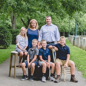 A family of 6 is posed on various stools and chairs on the paved path in Noblesville's Forest Park. They are coordinated in various shades of blue with the park trees softly focused in the background. Photo Credit: Kate Plummer - Studio Kate Portrait Design - Noblesville Family Photographer