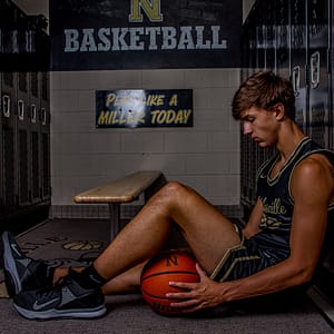 Noblesville Senior High School Graduate Cameron Karns is featured in the dark and moody locker room scene.  He’s sitting on the ground, leaned up against a locker, with a basketball under his knees, wearing his Millers jersey.  It’s a dark, quiet scene in which he looks down in great thought. Credit: Studio Kate Portrait Design - Noblesville Senior Pictures
