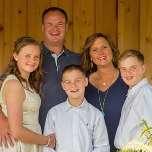 The Taylor family poses in front of a rustic knotty pine wall. The 3 teenagers are held gently by their parents with smiles all around. Photo Credit: Kate Plummer - Studio Kate Portrait Design - Noblesville Family Portrait Photographer