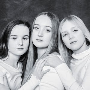3 sisters pose straight faced against a muddled grey studio backdrop in a classic formal black and white family portrait. The image is in a high-key studio style, which accentuates their young pale faces and white sweaters. Long straight hair flows between their heads with the younger outside girls tilted slightly toward the older teen in the center. Photo Credit: Kate Plummer - Studio Kate Portrait Design - Noblesville Family Portrait Photographer