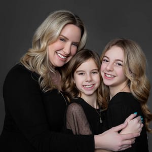 A mother and her two daughters embrace with the youngest in the middle. They are dressed in formal black outfits against a muddled grey studio backdrop creating a classic formal portrait. Photo Credit: Kate Plummer - Studio Kate Portrait Design - Noblesville Studio Portrait Photographer