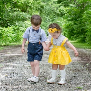 Two sibling toddlers hold hands as they focus on the gravel path they are walking down. The young boy is in blue shorts and suspenders with a matching bow tie. His younger sister is wearing a bright yellow dress with wide shoulder straps and a matching sunflower headband. Photo Credit: Kate Plummer - Studio Kate Portrait Design - Noblesville Family Portrait Photographer