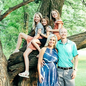 3 young girls are seating up on a large tree branch above their parents who are standing together in front of the tree. Photo Credit: Kate Plummer - Studio Kate Portrait Design - Noblesville Family Pictures Photographer
