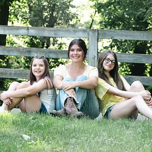 3 sisters are posed together, sitting in the grass in front of a weathered 3-rail wooden fence. This location can be found in Forest Park in Noblesville, just at the west end of the pedestrian bridge which crosses SR19 and leads right into downtown. Photo Credit: Kate Plummer - Studio Kate Portrait Design - Noblesville Family Portrait Photographer