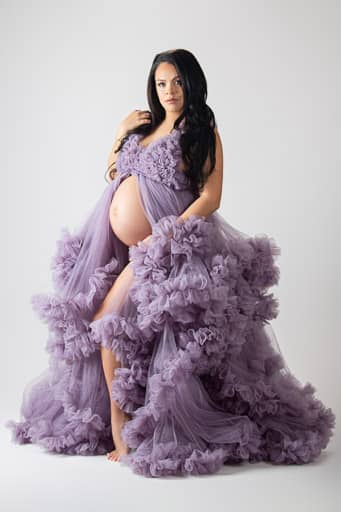 Maternity photo of a beautiful black haired pregnant woman in a big, fluffy, purple open-front dress, with her pregnant belly and left leg exposed exposed. Image courtesy of Studio Kate Portrait Design specializing in maternity pictures.