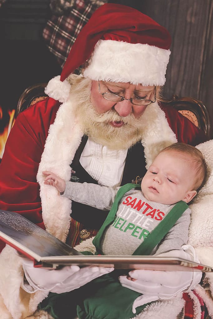 Santa Claus sits in his chair holding a small baby and reading The Night Before Christmas. Photographer Credit: Kate Plummer - Studio Kate Portrait Design - Noblesville Santa Experience