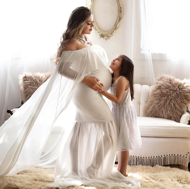 A pregnant mother is stands looking down and holding hands with her elementary aged daughter. This is studio pictures shows a living room like scene behind them while sunlight from the window shines through the long translucent white dress of the mother highlighting the beautiful curves of her maternal body.