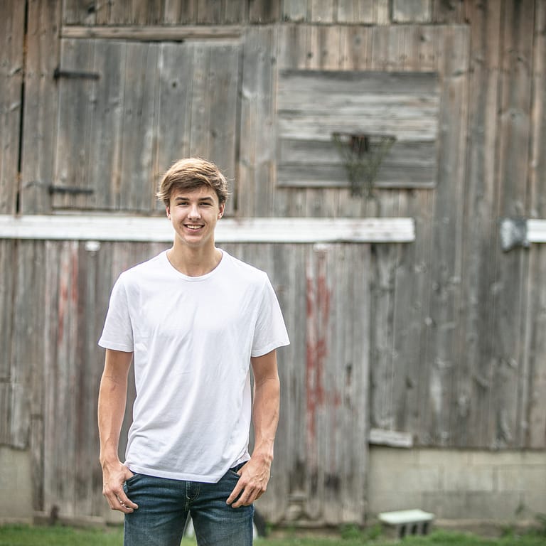 Noblesville High School basketball player Cameron Karns is in a simple white t-shirt standing in front of an old, weathered barn.  Just above the barn door is a similarly weathered backboard and rusty basketball rim. For years this has been a common sight in Indiana and looks like it could be a scene right out the movie “Hoosiers”. Credit: Studio Kate Portrait Design - Noblesville Senior Pictures