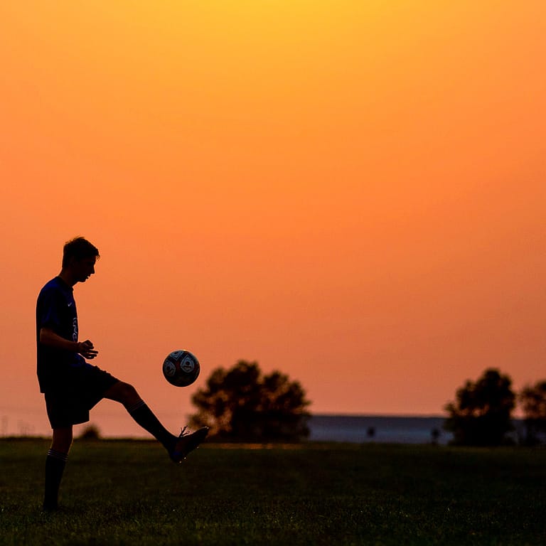 Noblesville Senior High School soccer player is just a black silhouette against a completely orange Autumn sky.  He’s kicking his soccer ball repeatedly, with nothing but the black outlines of trees along the bottom of the landscape. Credit: Studio Kate Portrait Design - Noblesville Senior Pictures