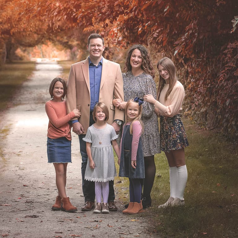 Mom, Dad, and 4 young girls are posed on a gravel trail with an amazing arch of red and orange autumn trees that follows the path behind them. The younger two sisters are standing directly in front of their parents, while the older two girls are holding their parents arms on each side of the group. Photo Credit: Kate Plummer - Studio Kate Portrait Design - Noblesville Family Portrait Photography