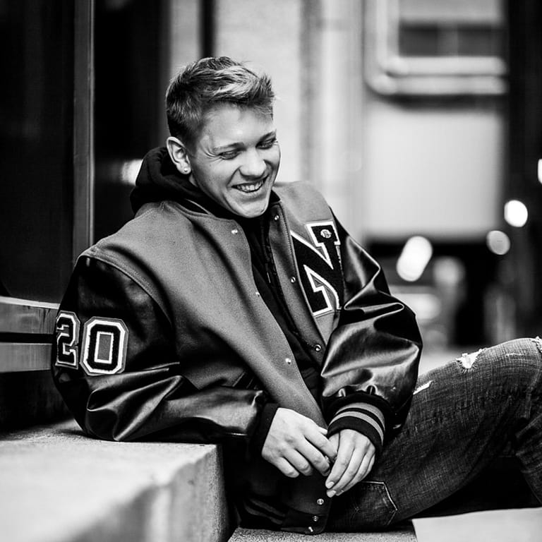 Noblesville Senior High School Student Chase is sitting on the steps downtown, wearing his NHS letter jacket with a big 2-0 on the sleeve, indicating that he graduated from high school in 2020.  His shy closed smile with his head bowed down presents a humble confidence. Credit: Studio Kate Portrait Design - Noblesville Senior Pictures