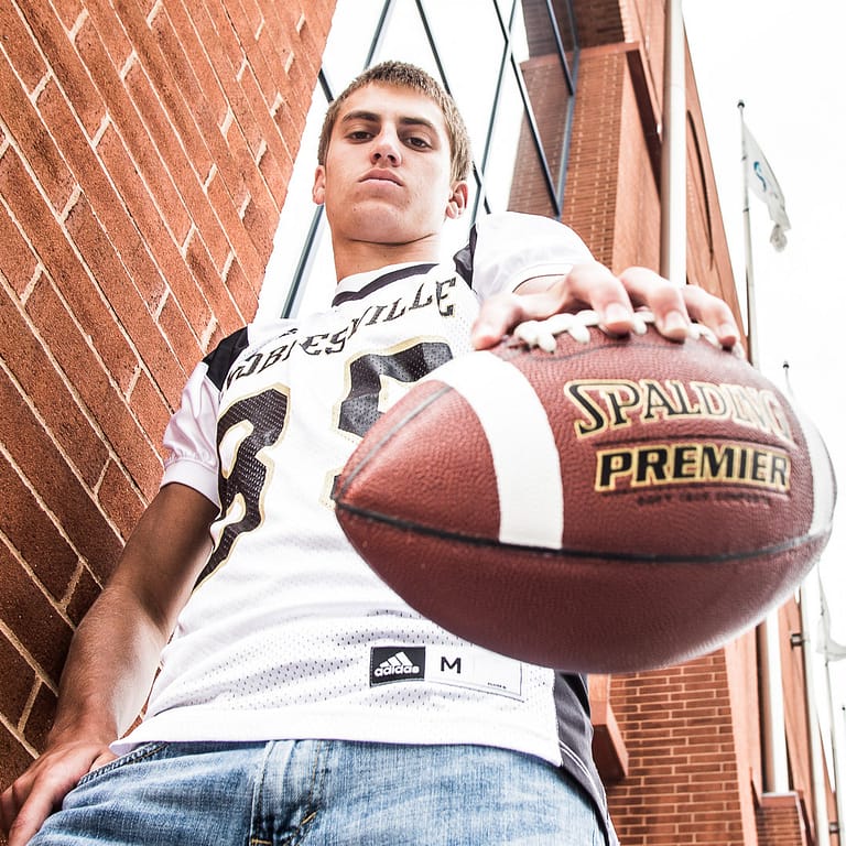 Noblesville Senior High School Student is standing in front of a towering brick building, palming a football and looking down sternly at the camera that is well below him.  This was shot with a wide angle lens which makes him appear to be a giant wearing his NHS Football jersey with jeans. Credit: Studio Kate Portrait Design - Noblesville Senior Pictures