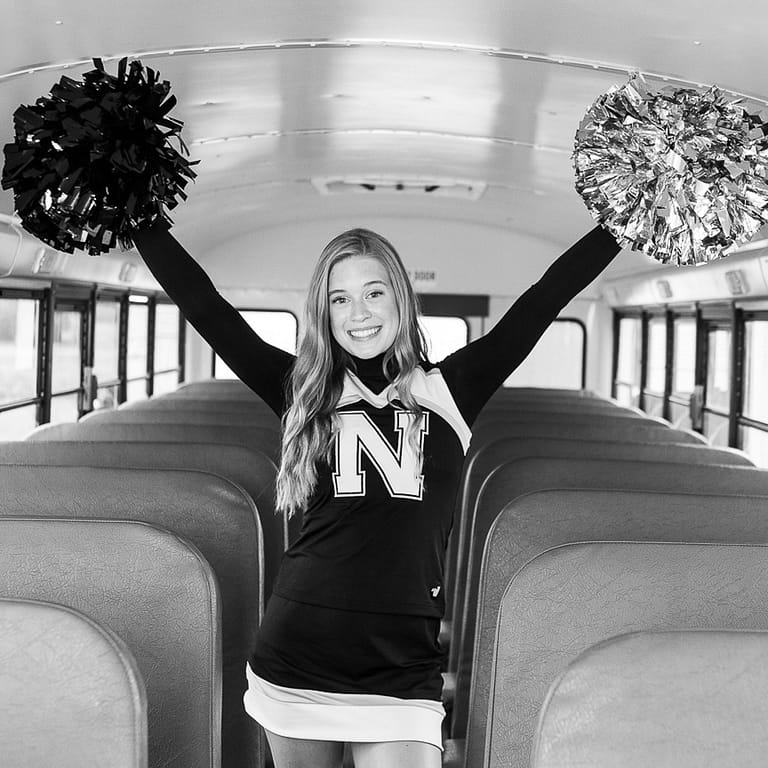 This Noblesville High School cheerleader is standing in the aisle of a school bus with pom poms in her hands and her arms stretched out and up.  There’s a big “N” that nearly covers the front of her cheer uniform, and her straight long blonde hair falls almost to her waist. Credit: Studio Kate Portrait Design - Noblesville Senior Pictures